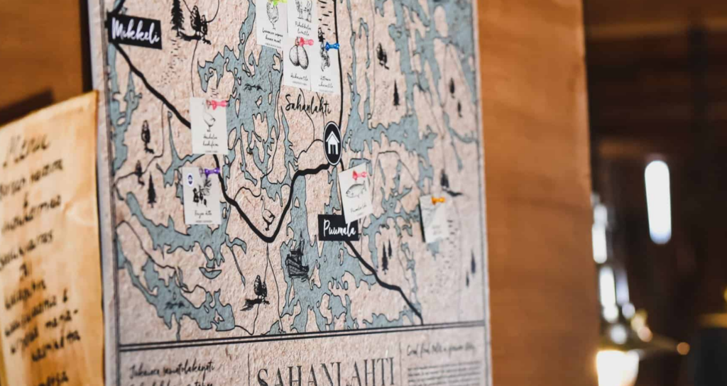 Seinällä vanhanaikainen piirretty, sinisen ja valkoisen sävyissä oleva Sahanlahden kartta, johon on kiinnitetty nastoilla lappuja. Lapuissa on kuvia lähiruoasta, kuten peruna ja kala, kiinnitettyinä kartalla siihen kohtaan, mistä ruoka tosiasiassa tulee.  