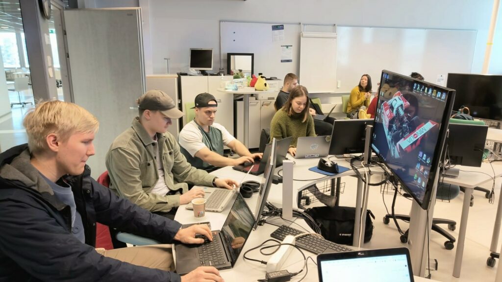 Kuvassa ryhmä opiskelijoita istuvat tietokoneineen ison yhteisen näytön edessä.